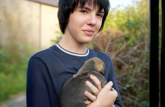 Teenage boy with black hair looking down with a gentle smile at a small newborn puppy sleeping peacefully in his arms outdoors