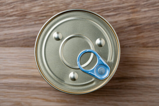 Top view closeup of sealed round aluminum tin can with pull tab mechanism, placed on textured brown wooden background. Simple food packaging mockup concept