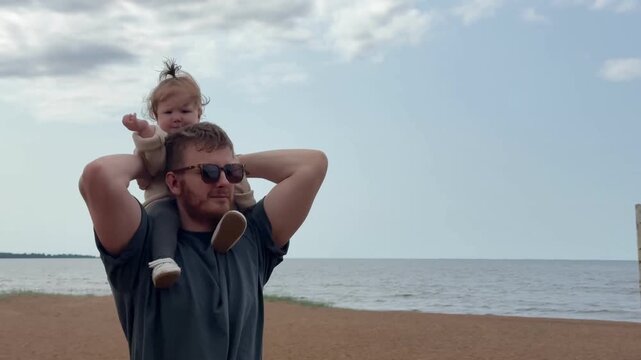 Adult man carries smiling toddler on shoulders beside calm shoreline with sea, sand, distant horizon, soft clouds, relaxed summer mood, and bright natural light