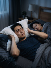Fototapeta premium Asian man husband covering ears with pillow while wife sleeps in dark bedroom. Sleep deprivation and relationship challenges vertical banner