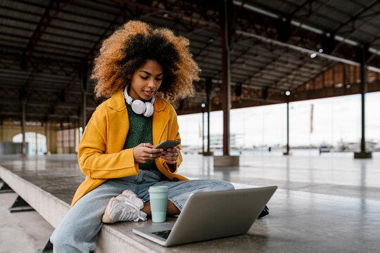 Afro woman using smart phone while sitting in front of laptop