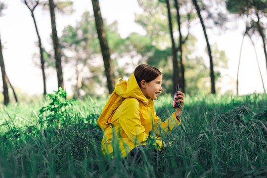 Girl with walkie-talkie wearing yellow raincoat and yellow backpack crouching on a meadow
