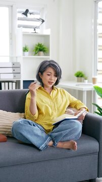asian woman 50s years old write down in notebook diary stay at home flat rest relax spend free spare time in living room .