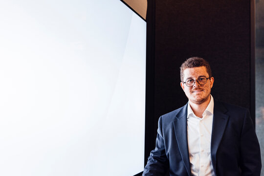 A smiling businessman stands by a blank screen in a modern office