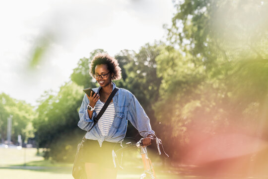 Young woman with cell phone pushing bicycle in park