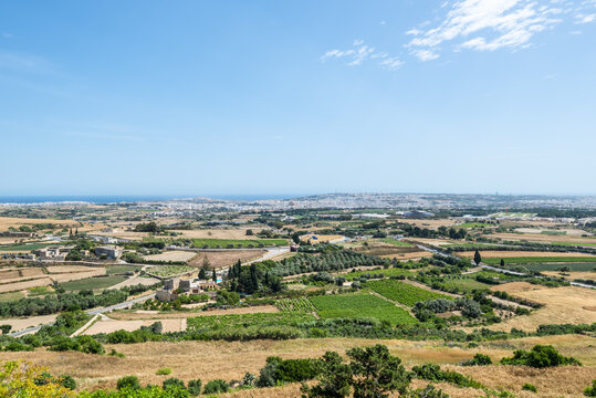Landscape views of central Malta from the fortified walls of Mdina, showing farmland, rural houses, green valleys and a distant coastal horizon under bright Mediterranean sunlight.