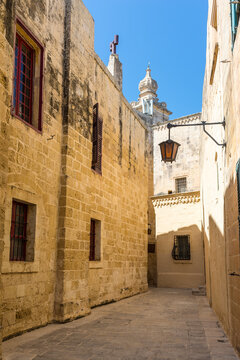A narrow limestone alley in the historic walled city of Mdina, Malta, with warm Mediterranean light revealing textured stone walls, shuttered windows and a distant dome rising above the old passageway