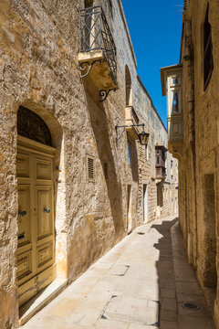 A charming narrow alley framed by golden limestone walls in the historic walled city of Mdina, Malta. Warm Mediterranean light and timeless medieval architecture create an inviting, atmospheric scene.