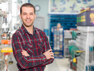 Obraz premium Portrait of a smiling male worker or store owner in a plaid shirt with crossed arms standing in a hardware store aisle next to a wall display rack full of wrenches.