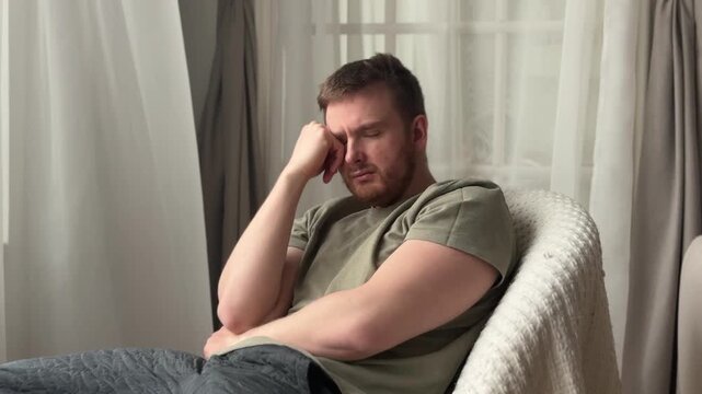 Fatigued depressed man reclining on plush beige sofa in calm living room with sheer curtains, knit pillow, soft ambient light, olive shirt, thoughtful mood and quiet home comfort