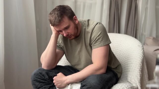 Contemplative man seated indoors with hand supporting chin, soft ambient light, neutral room tones, textured chair, relaxed posture, downward gaze, quiet reflective mood