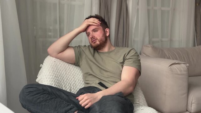 Fatigued depressed man reclining on plush beige sofa in calm living room with sheer curtains, knit pillow, soft ambient light, olive shirt, thoughtful mood and quiet home comfort