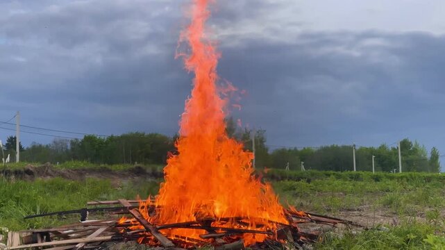 Rural bonfire roaring through piled timber and debris in open field beneath heavy clouds, sending tall orange flames, glowing embers, drifting smoke, and harsh heat across surrounding grass