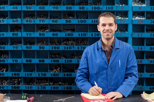 Portrait of a smiling male worker in a blue uniform writing on a notepad at a counter, with organized blue shelving bins full of metal parts in the background of a hardware store or warehouse.