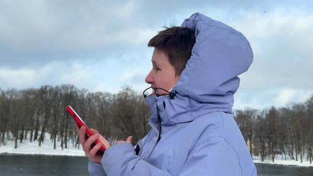 Side profile of person in lavender hooded winter jacket gazing at bright red smartphone beside snowy lakeshore with leafless trees, icy water, cloudy sky, and windy cold outdoor atmosphere