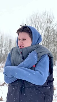 Young woman standing outdoors in snowy winter landscape with crossed arms, pursed lips and blue hooded jacket, facing cold weather with calm resilient stance, with snow, snowfall, winter, outdoors