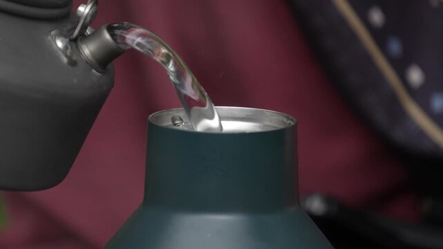 Close up of pouring fresh clear water from a camping kettle into a reusable thermal bottle flask outdoor. Eco friendly hydration concept.