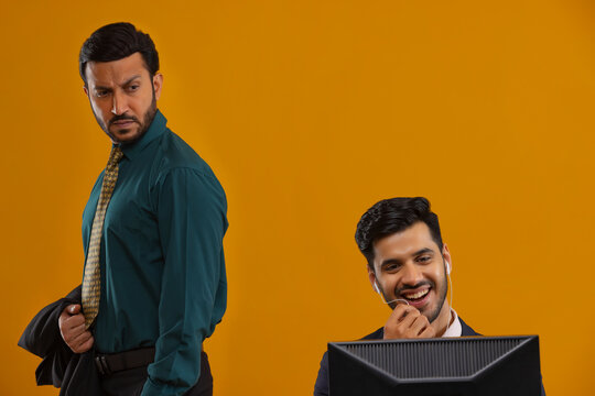 A corporate employee in formal clothing looking intently at colleague who is talking on earphones and working on laptop.
