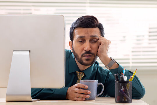 A corporate employee in formal clothing sitting behind desktop screen with drowsy eyes about to sleep.