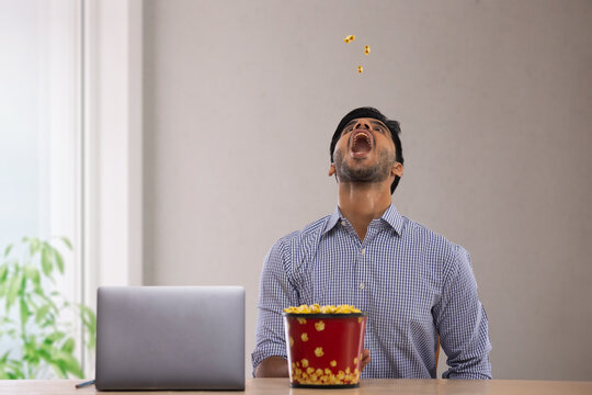 A  corporate employee in formal clothing sitting with a laptop catching popcorn with mouth.