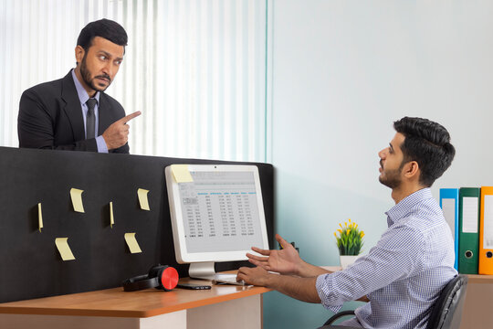 An angry boss in formal business suit scolding junior corporate employee at workdesk.