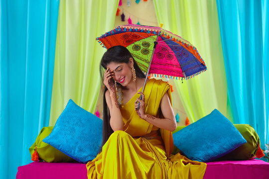 Indian bride in yellow saree looking down while sitting on stage with holding a umbrella over head