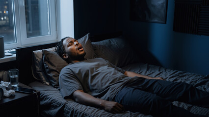 Fototapeta premium Black man patient lying on bed gasping for air in dark bedroom at night. Sleep apnea and sleep disorder awareness