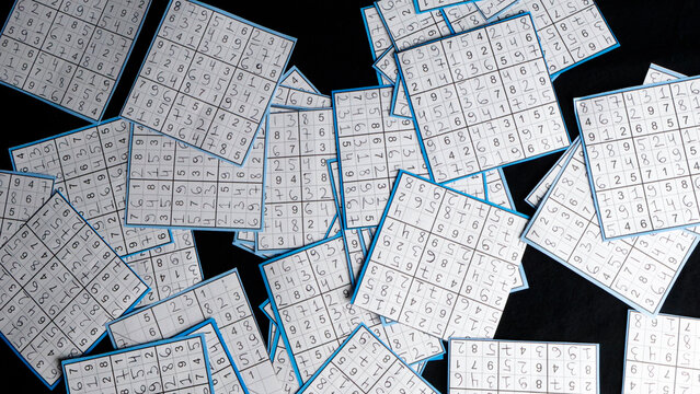 Many small paper squares with handwritten numbers forming completed sudoku puzzles lying arranged randomly on a dark studio background, representing concepts of problem solving and intelligence
