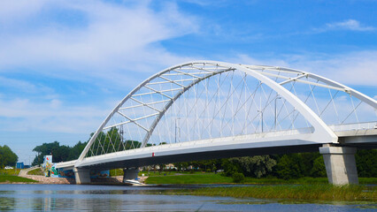 Fototapeta premium Modern white arch bridge connecting land over a reflective river with green banks, dense foliage, and a bright blue sky with wispy clouds during summer