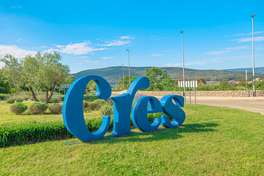Large blue Cres sign stands on a grassy area, a landmark welcoming visitors to the Croatian island with surrounding green hills and a bright summer sky of Cres Island in Croatia