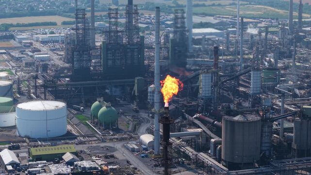 Tight close-up aerial shot tracking the thermal combustion flames of a refinery flare stack against a backdrop of industrial storage silos and processing units.