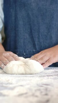 Vertical video: Floured hands shaping dough ball after resting at bench for proofing, denim apron