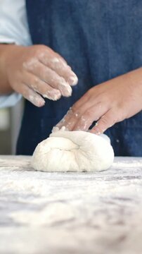 Vertical video: Woman in denim apron lifting dough, shaping dough into round to bake on floured top