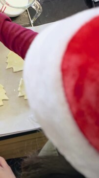 Vertical video: After cutting dough, mom in Santa hat helping child placing cookie cutouts on tray