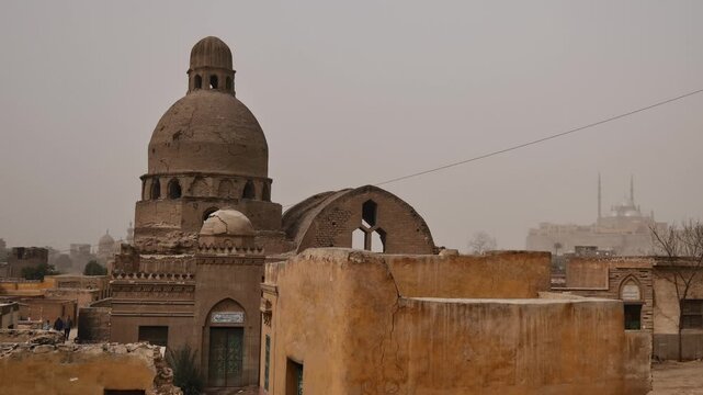 Moody view over Cairo necropolis, Egypt, Mamluk era mausoleum domes and crumbling tomb structures among palm trees with sandstorm casting orange haze over city.