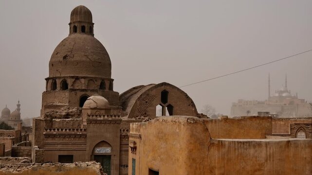 Dusty panoramic view over Cairo City of the Dead necropolis, Egypt, historic Islamic mausoleum domes rising above palm trees and crumbling brick tomb buildings in sandstorm haze.