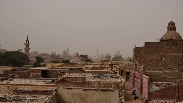 Medieval Islamic mausoleums and tomb structures of Cairo City of the Dead necropolis, Egypt, surrounded by palm trees and old buildings under orange dusty sky.