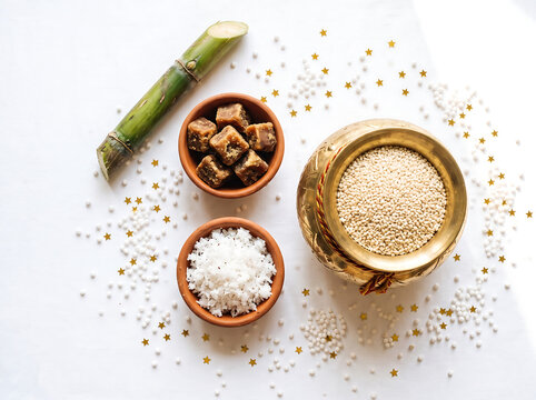 Top-down bright flatlay of a festive still life with a brass kalash of sesame seeds, a bowl of jaggery, and fresh sugarcane sticks on pure white background