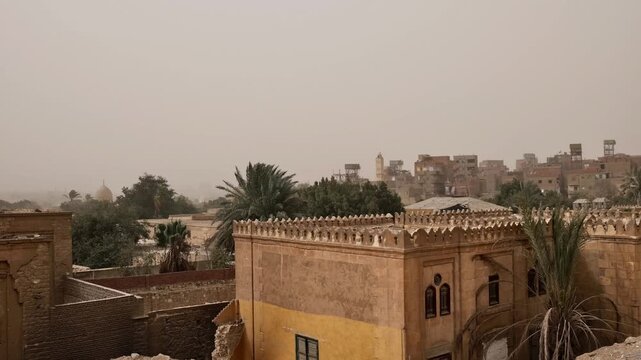 Medieval Islamic mausoleums and tomb structures of Cairo City of the Dead necropolis, Egypt, surrounded by palm trees and old buildings under orange dusty sky.