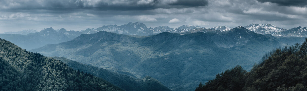 Layers of green and blue mountains extending into the distance under a vast, dark sky, with snow capped peaks