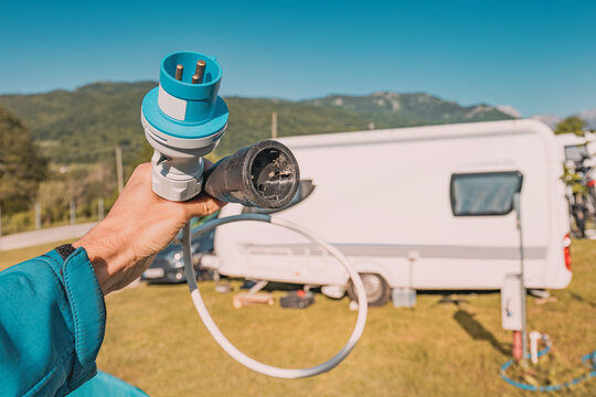 Hand holding electric hookup adapter cable at caravan campsite with mountains. Camping concept.