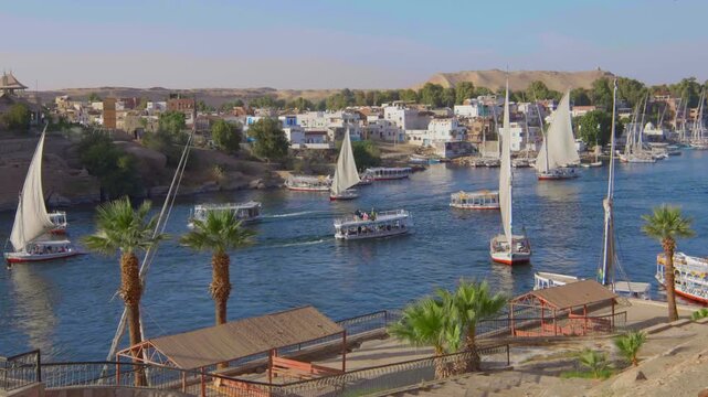 Traditional felucca sailboats and tourist boats on Nile River at Aswan, Egypt, with Elephantine Island Nubian village, palm trees and golden sand dunes behind. Timelapse