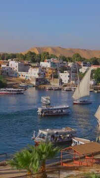 Scenic view of Nile River at Aswan, Egypt, with traditional felucca sailboats and tourist ferries, Elephantine Island village, palm trees and sandy desert hills in background. Timelapse