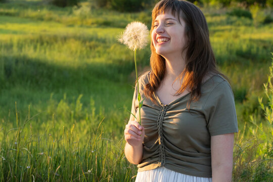 Happy young woman holding dandelion in summer meadow at sunset