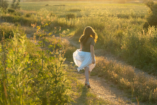 Young woman walking on a dirt path through a summer meadow at sunset