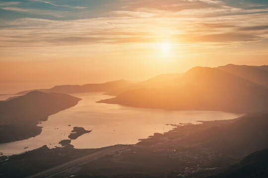 Bay of Kotor, Montenegro, bathed in golden light during sunset. The calm water reflects the vibrant sky with surrounding mountains