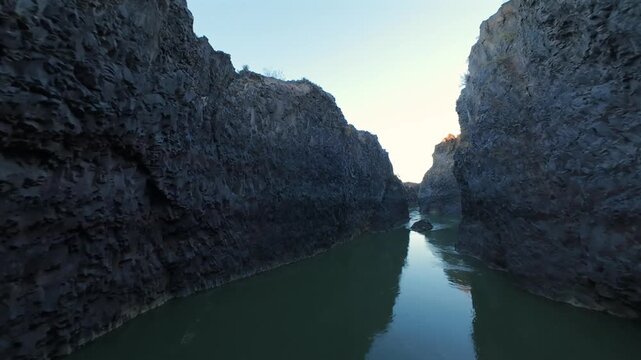 Dynamic FPV drone video soaring through a dramatic river canyon in La Payunia Reserve, Mendoza. Rugged volcanic cliffs, winding watercourse, golden hour light. Pristine nature aerial footage.