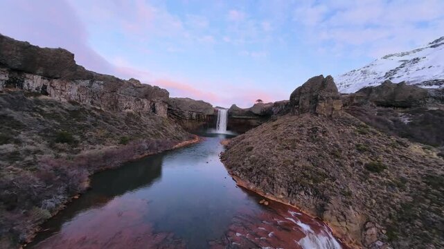 Salto del Agrio waterfall flows through the volcanic canyon of Caviahue Copahue, Neuquen, Argentina, revealing red mineral deposits, basalt cliffs and snowy peaks, FPV flyover.