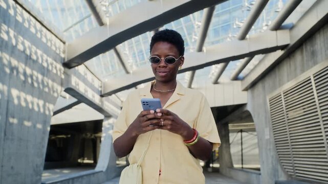 Tracking medium shot of smiling African American young adult woman using smartphone in modern urban setting. Footage is ideal for mobile application marketing and digital communication campaigns