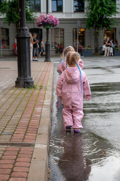Two children in pink rainwear walking through a large puddle on a wet city street